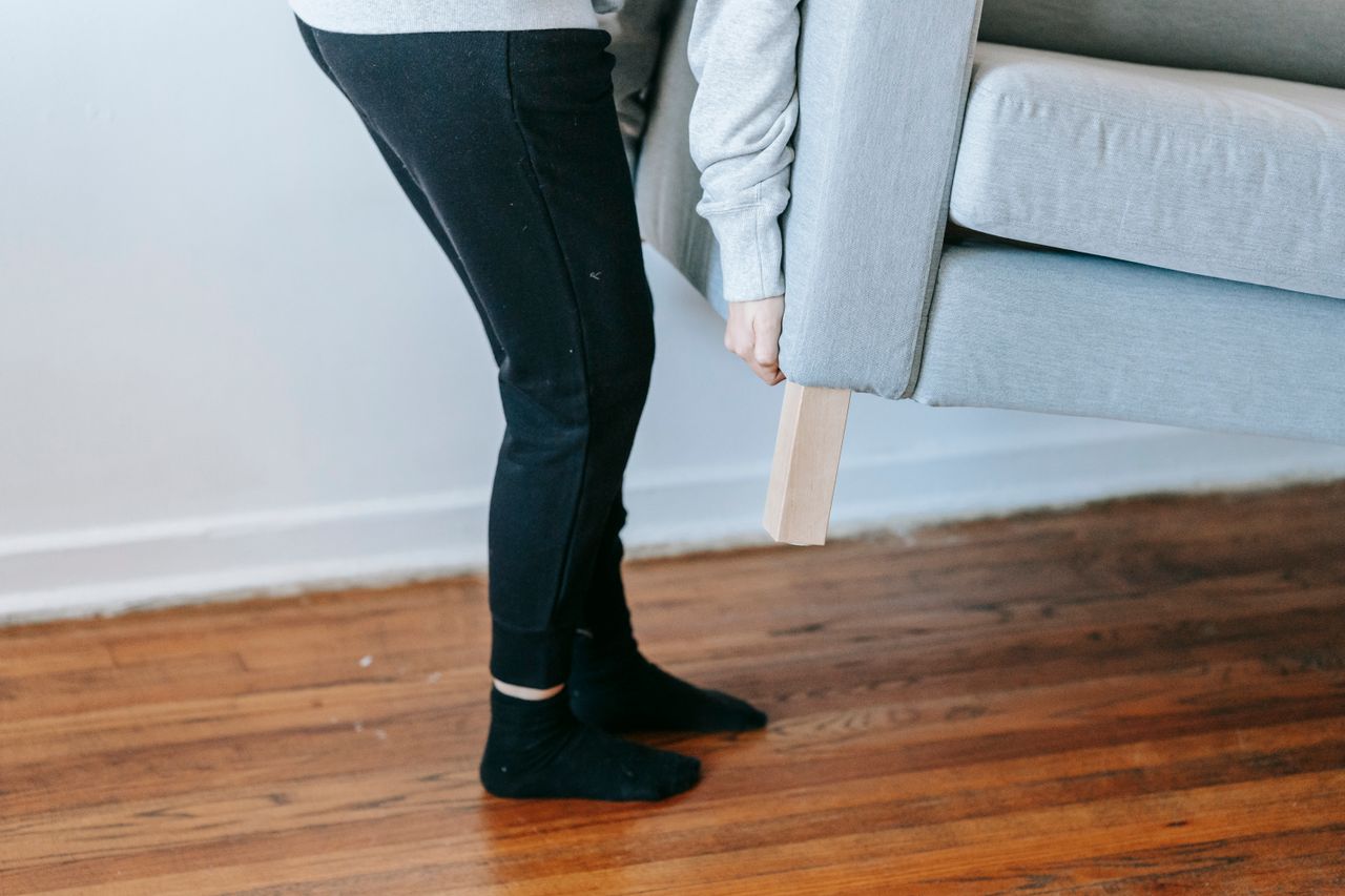 Une personne soulève un canapé dans un salon minimaliste aux sols en bois.